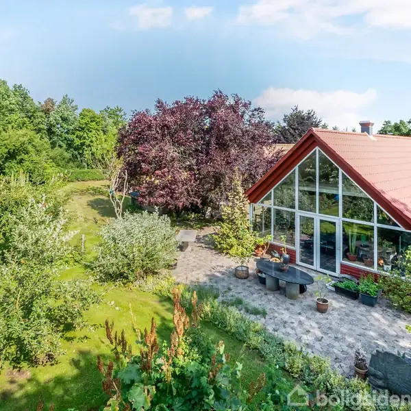 En moderne villa har en stor glasfacade mod en frodig have. Haven indeholder forskellige træer og buske, mens en terrasse foran huset har siddepladser og urtepotter.
