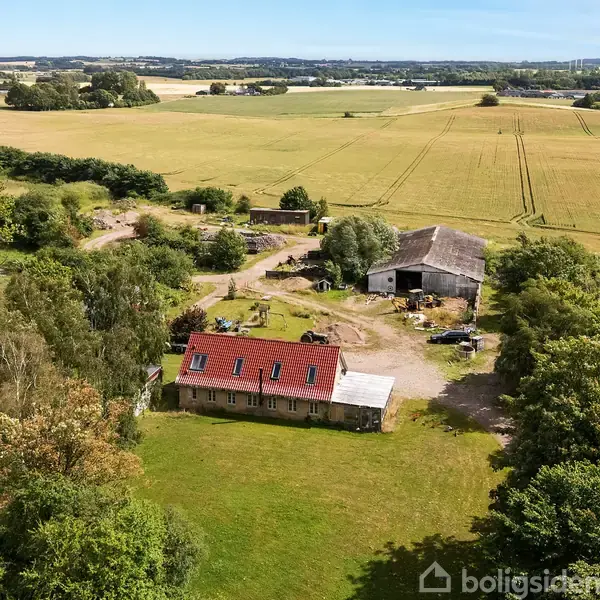 Et rødt taghus med vinduer står midt på en gård omgivet af åbne marker og træer. I baggrunden ses en lade og grønne landskaber strækker sig mod horisonten.