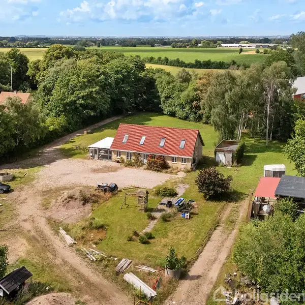 En landlig ejendom ses med et rødt tag, omgivet af grønne træer og marker. Foran huset er der en grusvej, biler og diverse landbrugsudstyr. Omkringliggende natur indrammer scenen.