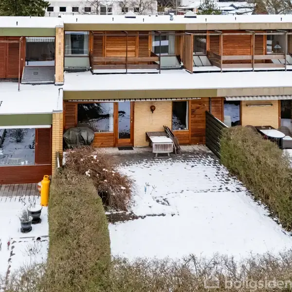 Rækkehus med snedækket terrasse og hæk i forgrunden. Ejendommen har store vinduespartier og træbeklædning på første sal. En gasflaske står ved siden af terrassen.
