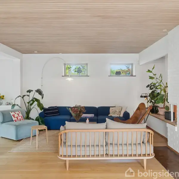 En moderne stue med en stor, blå sofa og lyse træmøbler omkring et sofabord. En pejs er indbygget i en hvid murstensvæg. Lyse trægulve og planter skaber en hyggelig atmosfære.