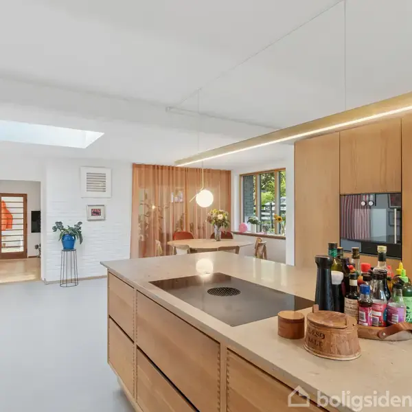 Køkken med træbordplade fyldt med flasker og krydderier, integreret vask og moderne inventar. I baggrunden ses spisestue med træbord og vinduer, der giver naturligt lys.