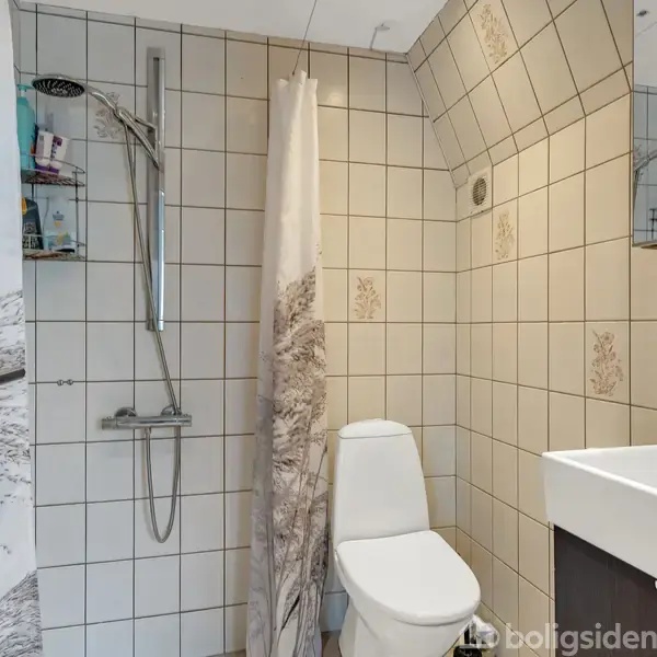 Bruser med integreret hylde ved siden af toilet i et flisebelagt badeværelse. Gardin adskiller bruseren. Håndvask med spejl hænger på højre væg. Trædør til venstre.
