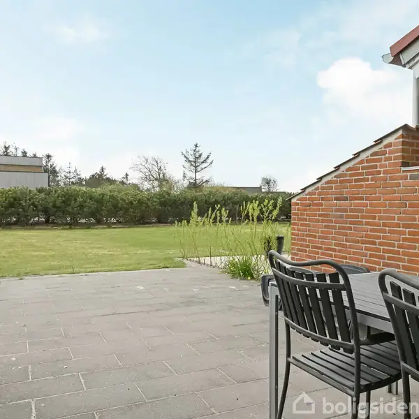 Terrassemøbler står på en flisebelagt terrasse ved siden af en murstensvæg. Græsplæne og buske omkranser området under en klar blå himmel.