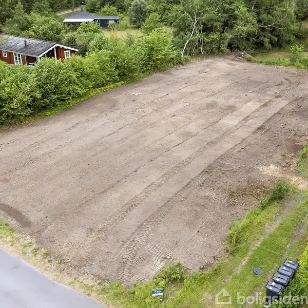 Jordlod ligger bar med traktorspor, omgivet af buske og træer. I baggrunden ses et rødt hus med gråt tag. To biler er parkeret ved en vej i nederste hjørne.