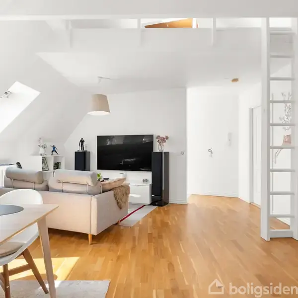 Stue med sofa og fladskærms-TV. En stige fører op til en hems. Store skråvinduer slipper lys ind. Et spisebord står nær sofaen. Lyse trægulve og hvide vægge.