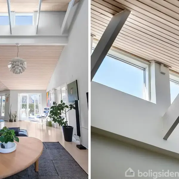 Stue med moderne indretning, trægulv og loft med ovenlysvinduer, der lader sollys strømme ind. I baggrunden ses et spiseområde og potteplanter, der bidrager til en lys, rummelig atmosfære.