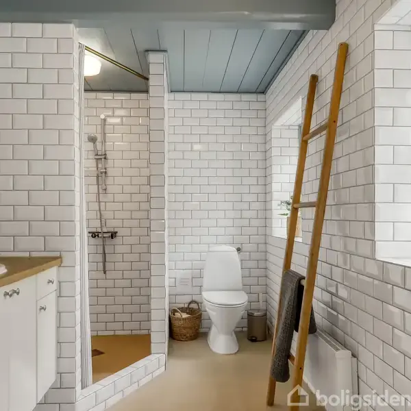 Badeværelse med hvidt flisebeklædning og trægulv. Bruseniche ved siden af et toilet, og en stige fungerer som håndklædeholder ved vinduet. Kurv og planter tilføjer indretningselementer.