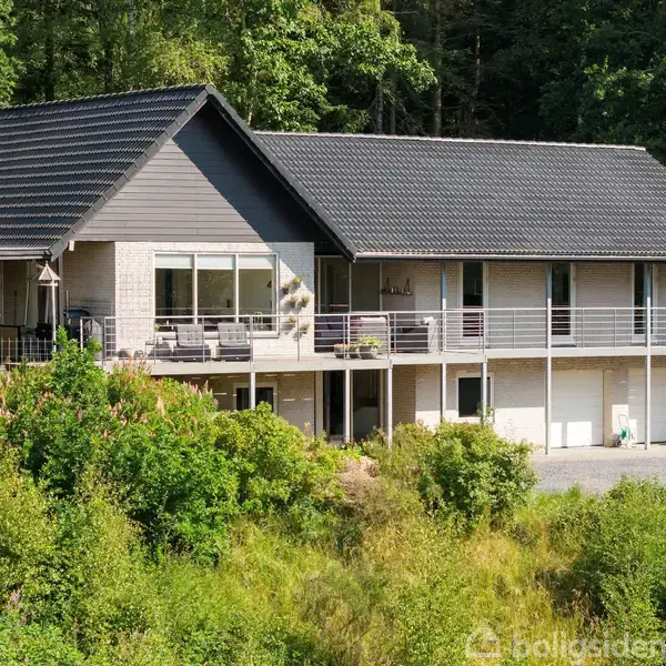 Et moderne hus med skråt tag står blandt frodig, grøn natur. Bygningen har store vinduer og en lang balkon. Træer omkranser ejendommen, som ligger i et skovklædt område.
