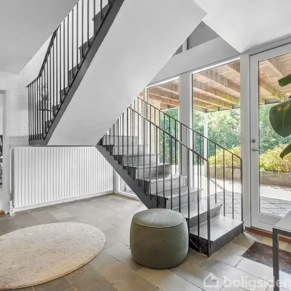 En trappe fører opad i en moderne entré med lys fra store vinduer. Et rundt tæppe ligger på gulvet nær en skammel, omgivet af en grøn plante og udendørsudsigt.