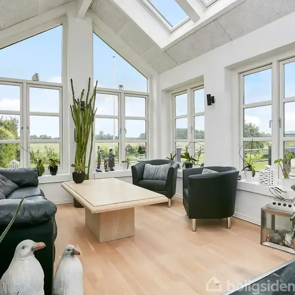 Sorte lædersofaer står i en vinterhave med store vinduer og et lyst træbord, omgivet af planter. Udsigt til en grøn have ses udenfor.