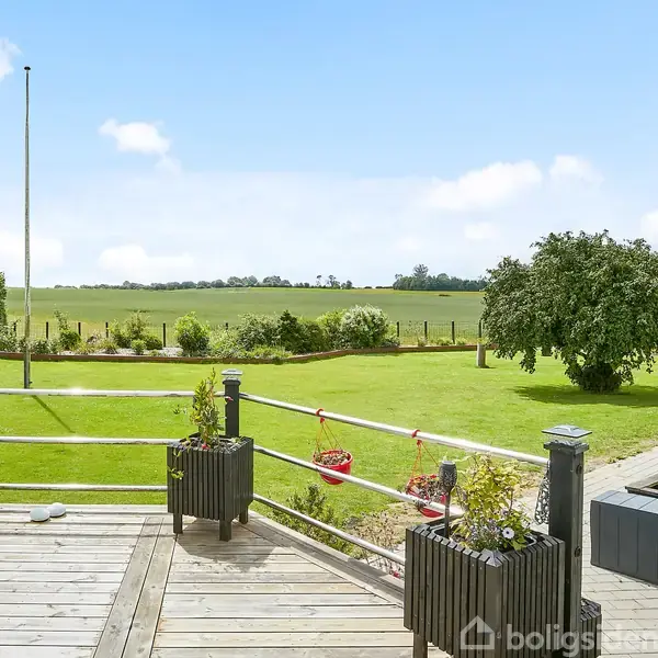 Terrasse med blomsterkasser står stille foran en velplejet græsplæne med et enkelt træ. Baggrunden viser åbne marker under en blå himmel med få skyer.