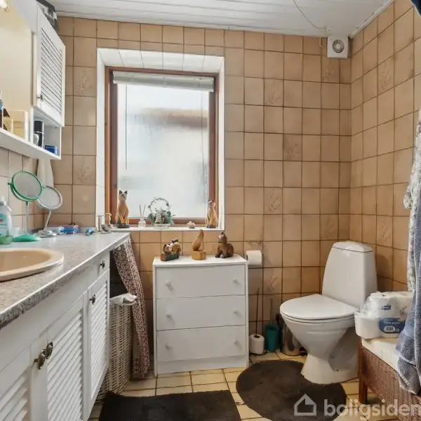 Badeværelse med brunlige fliser. Vask med spejl på venstre side og toilet til højre. Et vindue midt i rummet og gulvmåtter på det ternede gulv.
