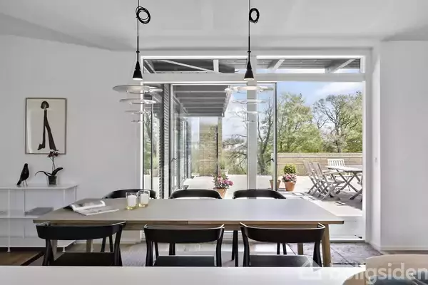 Spisebord med sorte stole står i et lyst rum. Store vinduer åbner ud til en terrasse med havebord og træer i baggrunden. Kunstværk og vase på en opbevaringsenhed.