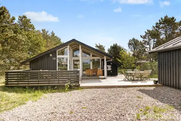 Sort sommerhus med store vinduer har nummeret "18"; en terrasse med havemøbler er omkranset af skov. Grusbelægning fører op til huset, der ligger i et naturrigt område.