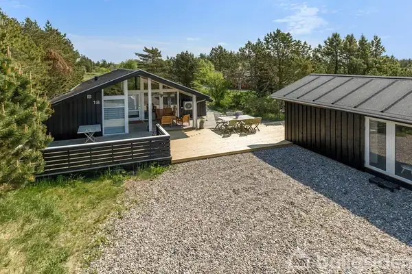 Et sort træhus med store glaspartier står i en skovklædt have. En grusbelagt indkørsel fører forbi et mindre anneks. Nummer '16' er synligt på huset.