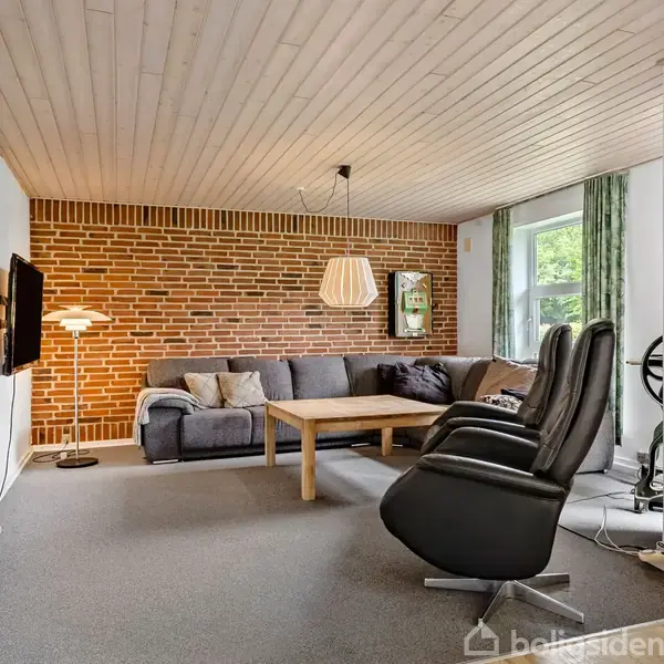 Stue med gråt hjørnesofaarrangement, der står foran en rød murstensvæg. To sorte lænestole står i forgrunden. Et stort vindue med grønne gardiner til højre. Fjernsyn hænger på væggen.