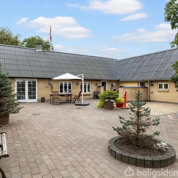 Et gårdhaveområde med flisebelægning omgivet af gulstensbygninger. Et bord med parasol står i midten, omgivet af planter i krukker og en bænk. Træer grænser op til området.