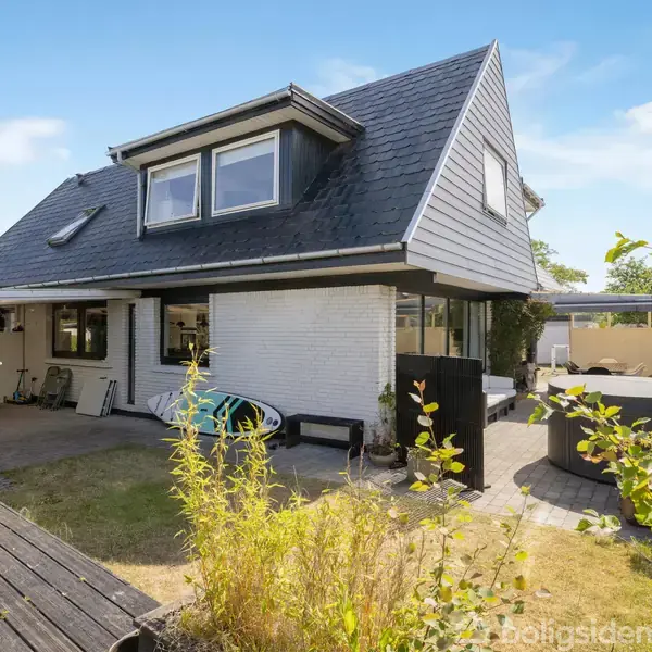 Hus med sort tag og to vinduer, omkranset af terrasse og have. Der er udendørs møbler og frodige planter omkring, hvilket skaber en afslappet atmosfære.