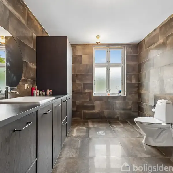 Rundt spejl hænger over en mørk vask i et moderne badeværelse. Toilet står ved siden af i et rum med store flisevægge og gulv, belyst af naturligt lys fra vinduet.