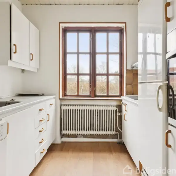 Et lyst køkken med hvide skabe står tomt. Et stort vindue med udsigt til træer giver naturligt lys, mens en radiator er placeret under vindueskarmen. Kokken er indrettet med ovn og vask.