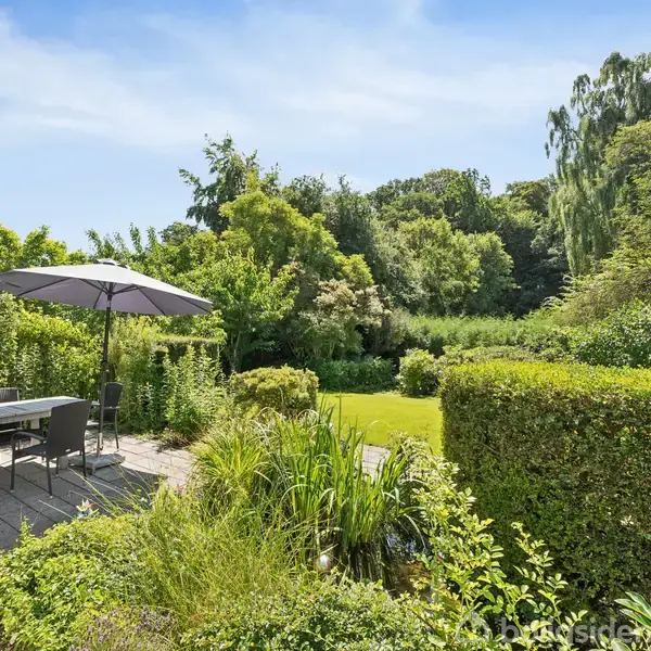 Terrasse med spisebord og stole under to store parasoller. Omkring terrassen er der frodig vegetation og trimmede buske, der grænser op til en stor græsplæne med omgivende træer.