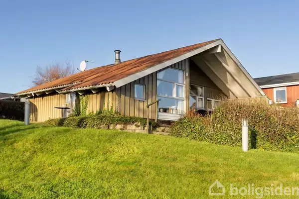 Et træhus med rødt tag står på en græsbeklædt bakke. Store vinduer på facaden reflekterer sollys, og en parabolantenne er placeret på taget.