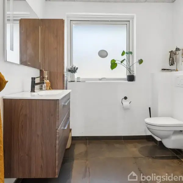 Et moderne badeværelse med væghængt toilet og træskabsarrangement. Et gult håndklæde hænger fra en krog. Et lille vindue med en plante står over toilettet.