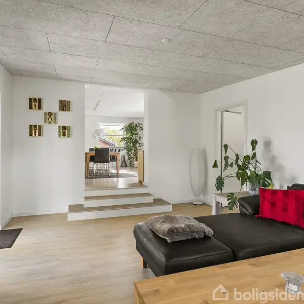 Sort sofa med røde puder står stille i en lys stue med plankegulve, indrammet af en glasdør til venstre og et spiseområde trin op i baggrunden.