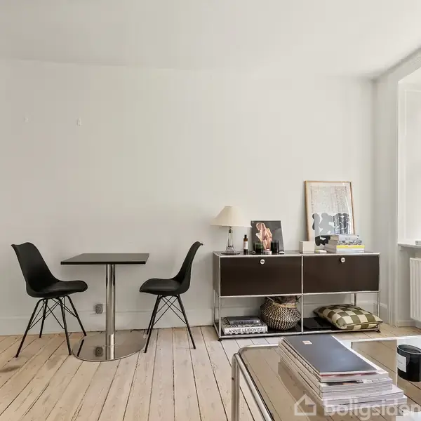 Et moderne rum har et lille, sort bord med to stole. Ved siden af står en skænk med bøger og en lampe. Store vinduer tillader naturligt lys at komme ind.