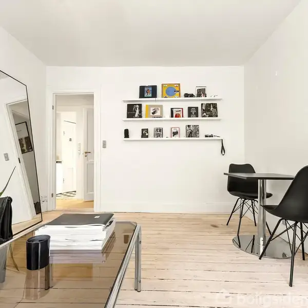 Stue med sort sofa og glasbord foran stor skråspejle på gulvet. På væggen hænger hylder med bøger og billeder. Rundt spisebord med sorte stole. Trægulv og hvidmalede vægge.