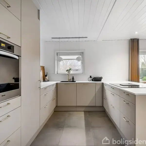 Et moderne køkken med hvide skabe og integreret ovn står stille i et lyst, ryddeligt rum. Store vinduer til venstre og i midten tillader naturligt lys at strømme ind.