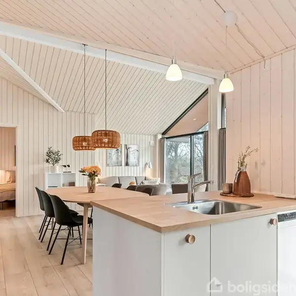 Køkkenø med vask står i et lyst, moderne rum. Spisebord med sorte stole nær vinduer, der giver udsigt til udendørs omgivelser. Loftet er højt med træpaneler, belysning hænger ned.