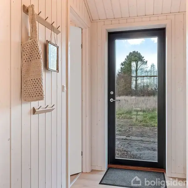 Glasdør åbner ud mod en have med træer i baggrunden. Indgang har lyse trævægge, en hvid dør til højre, en knagerække og hængende taske til venstre.