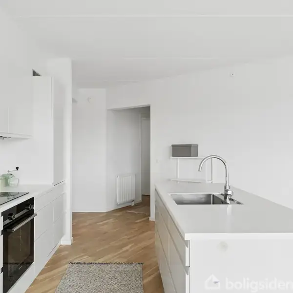Hvidt, moderne køkken med en stor køkkenø udstyret med en vask. Ovnen er indbygget i et skab til venstre, omgivet af en lys, minimalistisk indretning på et trægulv.