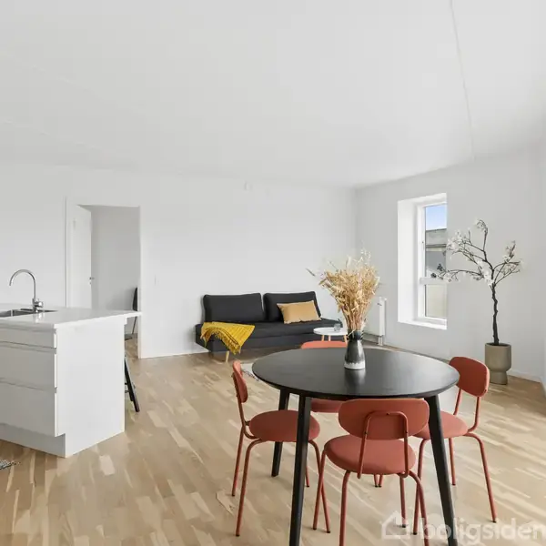 En rund spisebord med røde stole står i et lyst, moderne køkken-alrum. I baggrunden ses en sort sofa, en gul plaid og en plante ved siden af store vinduer.