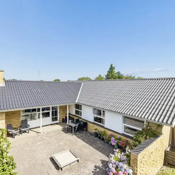Et hus med mørkt tag og gule mursten. Det står stille i en have med terrasse og blomsterbede, omgivet af grønne buske og et stakit under en klar blå himmel.