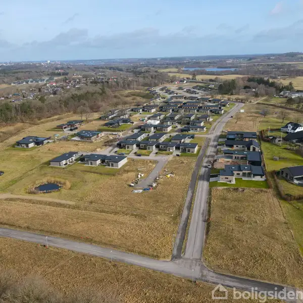 Boligområde viser række af moderne huse under konstruktion og færdige huse, omkranset af åbne marker og skove. Areaet struktureres af veje og inkluderer små vandhuller.