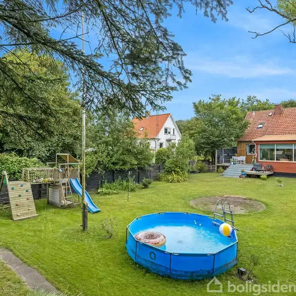 En rund, blå swimmingpool står midt i en grøn have. Omkring er der en rutsjebane, legeredskaber og en rød bygning. Himlen er blå, og træer omgiver området.