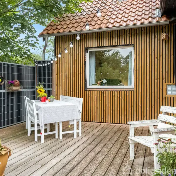 Træterrasse med hvidt bord og stole under en snor af lyskæder. Vægge af trælameller og mursten, omkranset af planter. En krukke med solsikker står på bordet.