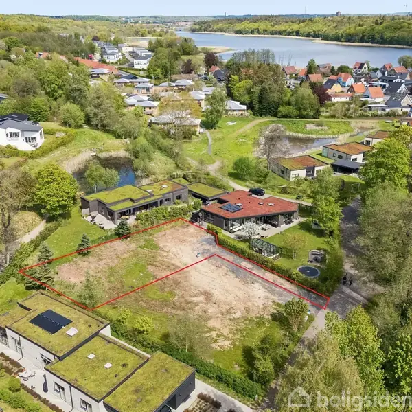 Grønt område ligger omkranset af beboelseshuse og tæt på en sø, afgrænset af en rød linje. Baggrunden består af flere huse og træer spredt i et naturskønt landskab.