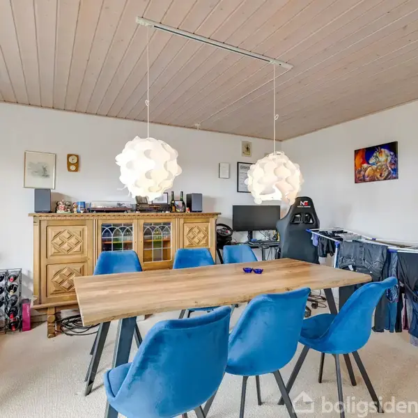 Et spisebord med seks blå stole står i et rum. To unikke hængelamper er over bordet. Omkring er der en vasketøjsstativ, et skab, og dekorationer på væggene.