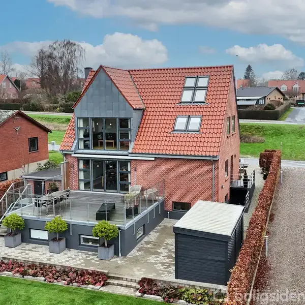 Et rødt murstenshus med en stor terrasse, beliggende i en forstad med flere lignende huse og en vej i nærheden. Hækken omkranser ejendommen.