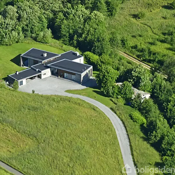En moderne bolig med fladt tag, beliggende isoleret omgivet af grønne områder og træer, set fra luften. En snoet vej fører op til boligens indkørsel.