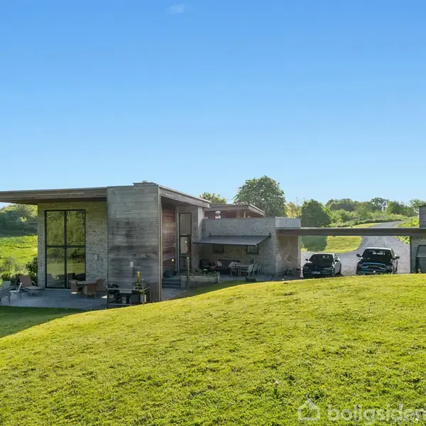 Moderne hus med fladt tag står på en grøn bakke. Ved siden af huset holder to biler parkeret på en asfalteret indkørsel. Omgivelserne er frodige med træer i baggrunden.