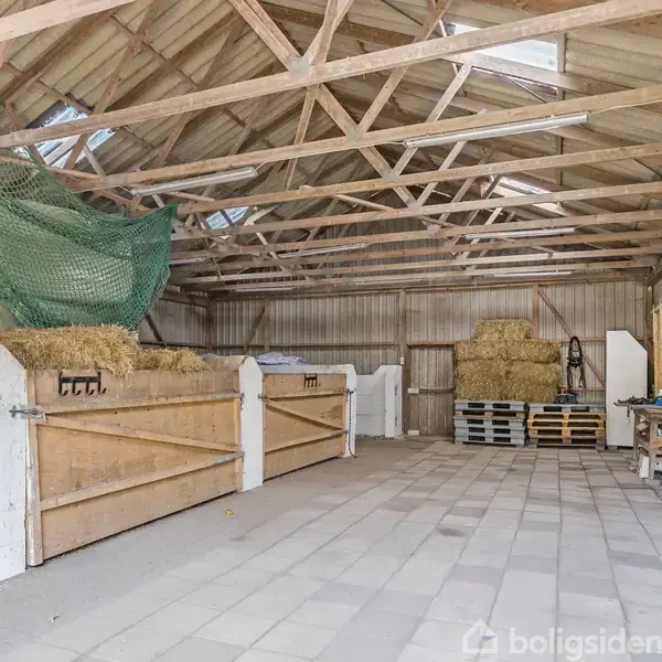 En lade med høballer stablet på båsene. Der er et stort grønt net hængende over dem. Rummet har synlige loftsbjælker og betonflisegulv. Der er paller og udstyr på bagvæggen.