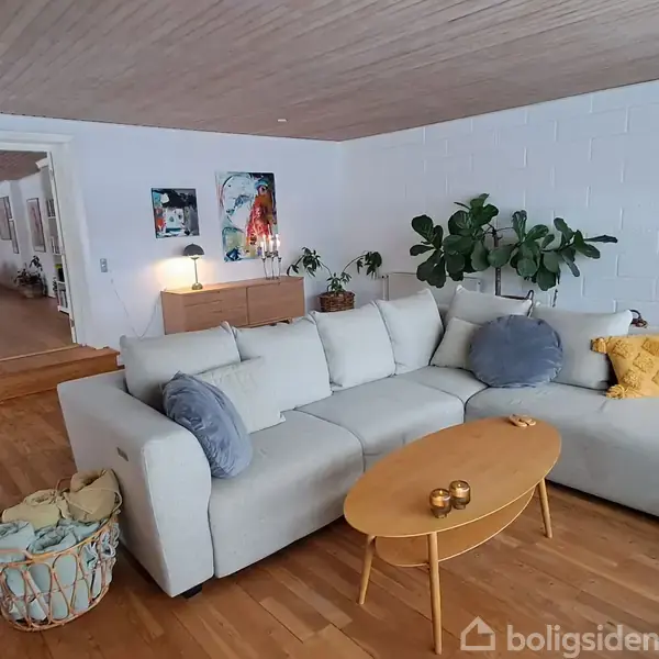 En stor, lys sofa med grå puder er placeret ved et rundt træbord med dekorative genstande. Rummet har trælofter, planter og kunst på væggene.