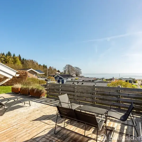 En træterrasse med havemøbler står i solen. Omgivet af en hæk og træer, med udsigt over et kystområde med huse og havet i baggrunden under en skyfri himmel.