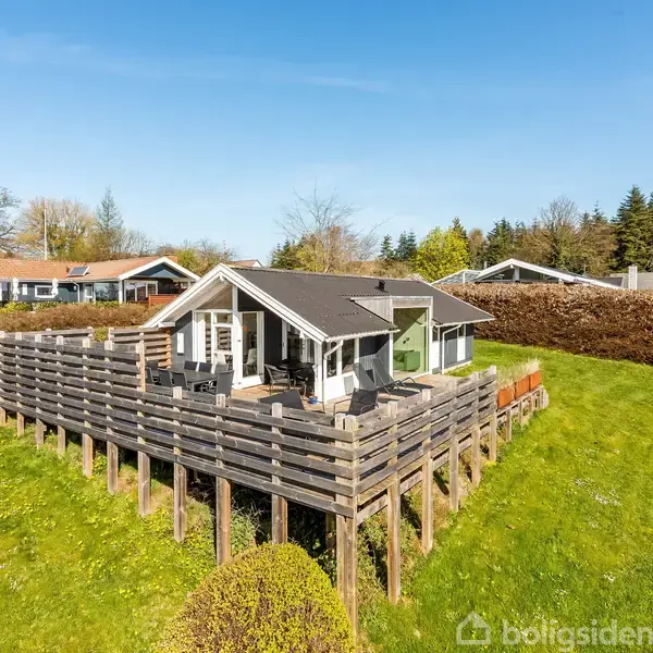 En lille træhytte står på en forhøjet træterrasse; omgivet af grønt græs og hække under en klar, blå himmel, i et villakvarter med andre huse i baggrunden.