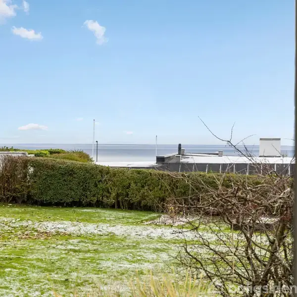 Fodboldmål står på en græsplæne med lidt sne, mens baggrunden viser en hæk, havudsigt og blå himmel.